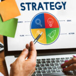 Persona señalando la pantalla de un portátil que muestra un diagrama circular colorido dividido en cuatro secciones etiquetadas como 'RESEARCH', 'IDEAS', 'ACTION' y 'PLAN'. Encima del diagrama aparece la palabra 'STRATEGY'. Sobre el escritorio hay una calculadora, papeles y notas adhesivas.