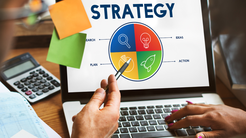 Persona señalando la pantalla de un portátil que muestra un diagrama circular colorido dividido en cuatro secciones etiquetadas como 'RESEARCH', 'IDEAS', 'ACTION' y 'PLAN'. Encima del diagrama aparece la palabra 'STRATEGY'. Sobre el escritorio hay una calculadora, papeles y notas adhesivas.